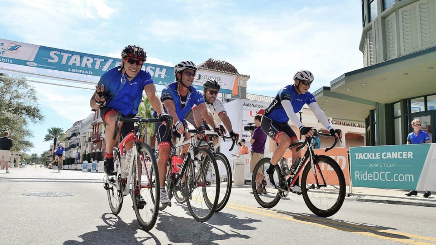 A group of cyclists wearing blue jerseys rides together past the start/finish line at a Miami Dolphins Challenge Cancer (DCC) event.